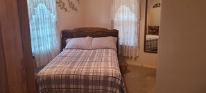 A bedroom with a bed, curtains, a wooden headboard, a mirror, and a carpeted floor.