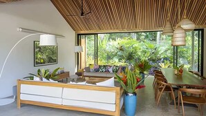 A modern interior with a wooden ceiling, a dining area with a table and chairs, a sofa, and a large window offering a view of greenery. - Caribbean Courtyard | Luxury Jungle Villa w Pool (Puerto Viejo de Talamanca)