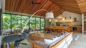 A spacious living area with a wooden ceiling, large windows, and a kitchen in the background. - Caribbean Courtyard | Luxury Jungle Villa w Pool (Puerto Viejo de Talamanca)
