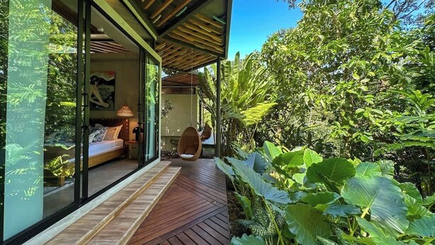A modern house with a wooden deck, a glass-enclosed bedroom, and lush greenery. - Caribbean Courtyard | Luxury Jungle Villa w Pool (Puerto Viejo de Talamanca)