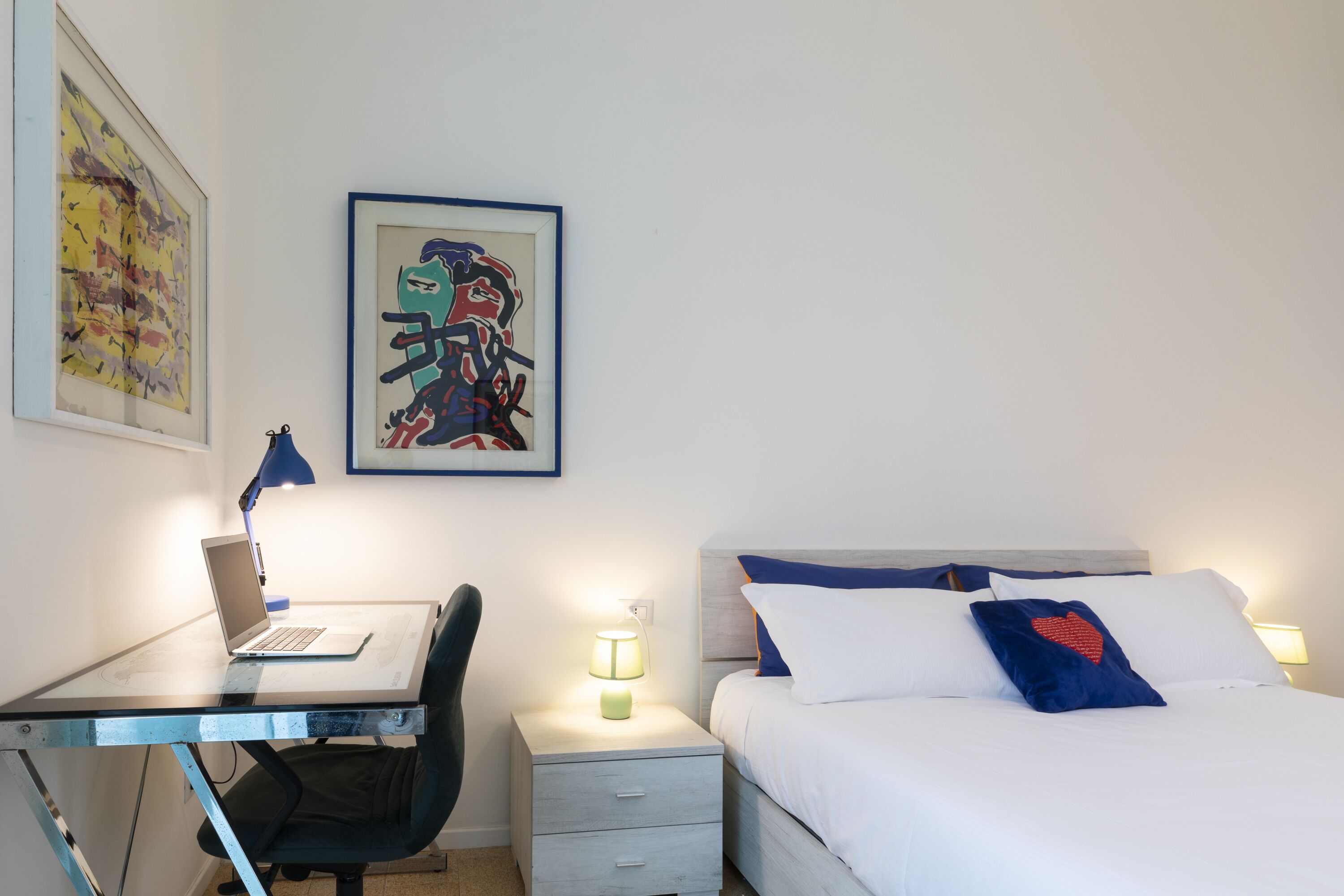 A hotel room with a desk, chair, laptop, bedside table with lamp, bed with pillows, and artwork on the wall.