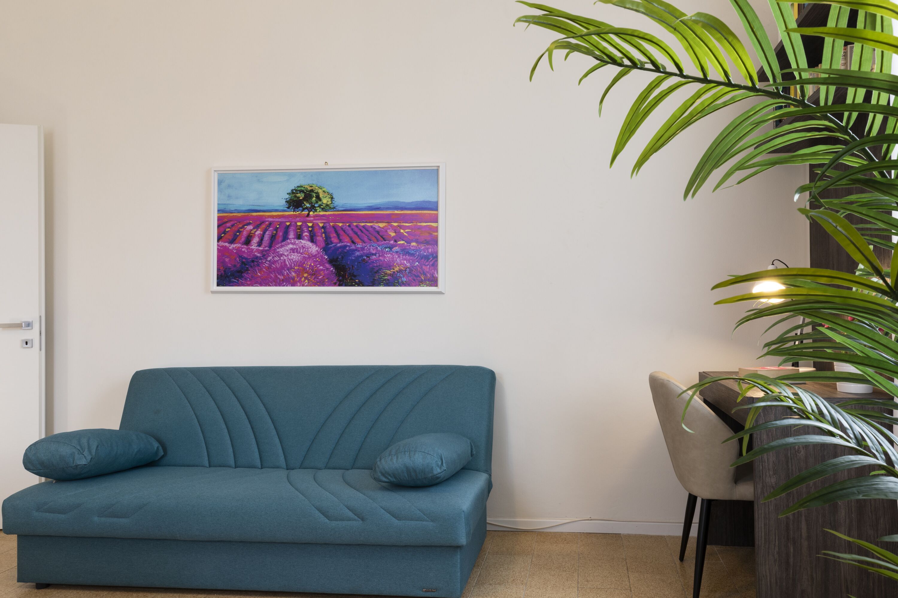 A teal sofa with two cushions, a framed painting of a lavender field, and a potted plant.