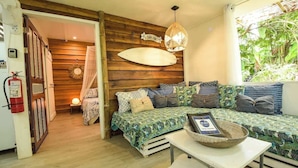 A cozy room with a bed, a table with a basket, a surfboard decoration, and a fire extinguisher. - Mini Up House · Romantic Caribbean Escape for 2 (Puerto Viejo de Talamanca)