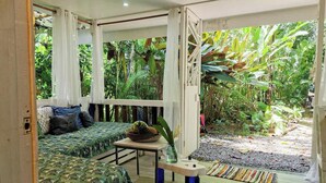 A room with a bed, a small table, and a view of a lush garden. - Mini Up House · Romantic Caribbean Escape for 2 (Puerto Viejo de Talamanca)
