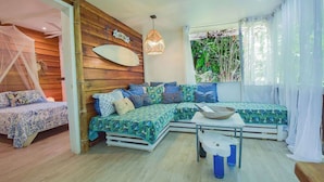 A cozy room with a wooden wall, a bed with patterned bedding, a sofa with cushions, a hanging light fixture, and a window with curtains. - Mini Up House · Romantic Caribbean Escape for 2 (Puerto Viejo de Talamanca)