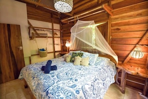 Une chambre dans une cabane en bois, avec un lit, une corbeille suspendue, une table avec une lampe et une moustiquaire.