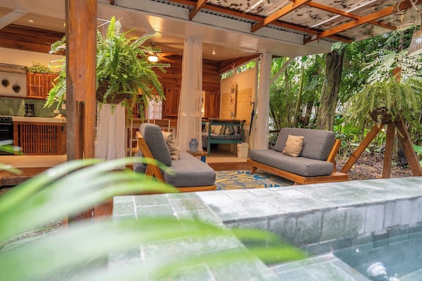 A cozy indoor space with a wooden ceiling, a seating area with a sofa and chairs, and a kitchen area with a stove and cabinets.
