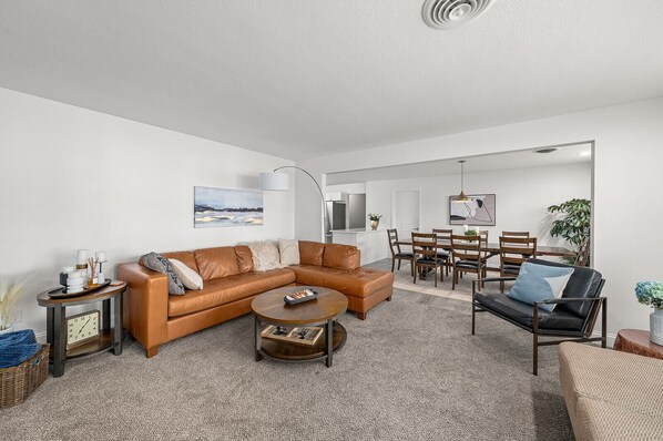 A modern living room with a brown leather sectional sofa, a round coffee table, and a black sofa with a blue pillow. There is a dining area with wooden chairs and a table, and a large mirror on the wall.