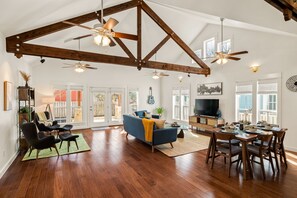 A spacious living room with a high ceiling, wooden beams, and a dining area with a table set for four. - Historic Dripping Springs-Clean, Quiet Designer Cottage Walk To Shops And Dining (Dripping Springs)