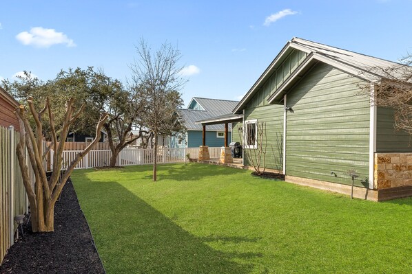 A residential property with a well-maintained lawn, a wooden fence, and a green building with a sloped roof. - Historic Dripping Springs-Clean, Quiet Designer Cottage Walk To Shops And Dining (Dripping Springs)