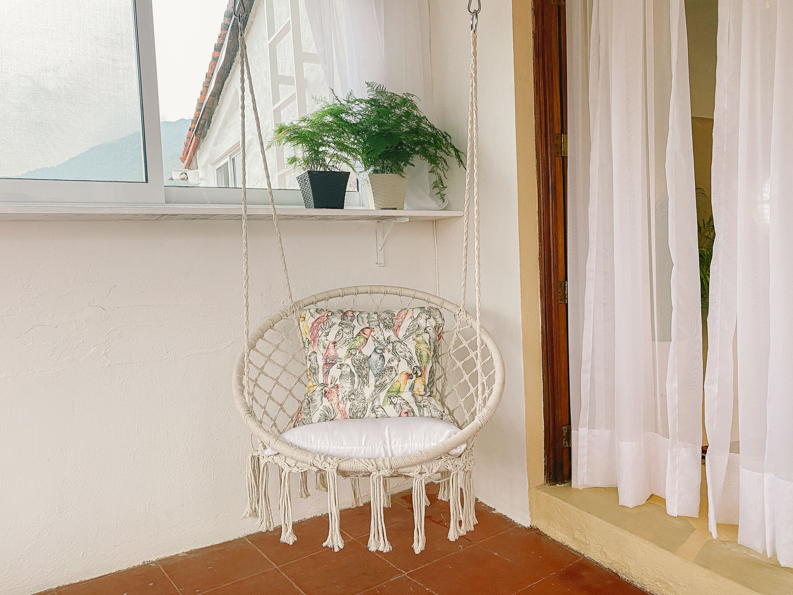 Una poltrona amaca in macramè con un cuscino, una pianta in vaso su uno scaffale e una finestra con tende.
