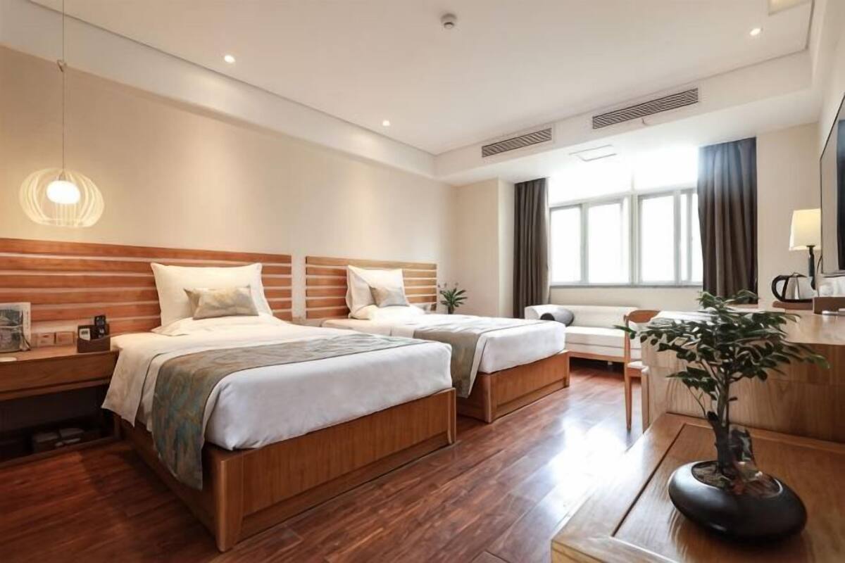 Een hotelkamer met twee bedden, een bureau, een stoel, een plant en een groot raam.