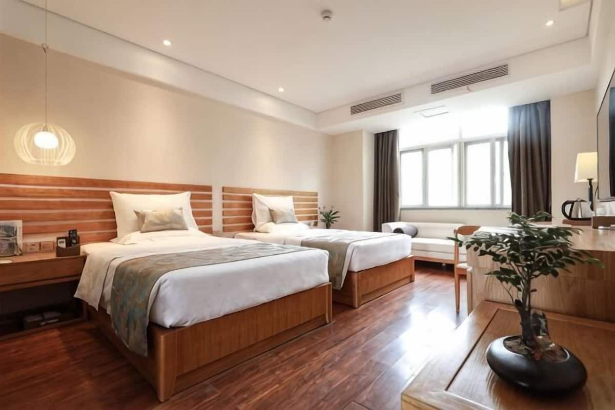 A hotel room with two beds, a desk, a chair, a plant, and a large window.