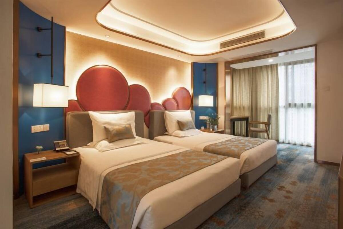 Een hotelkamer met twee bedden, een bureau, een stoel, een lamp en een groot raam met gordijnen.