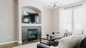 A modern living room with a fireplace, a flat-screen TV mounted above it, and a chandelier. There are two white armchairs and a small table with a globe and books. - Spacious 3 BR House w/hot tub in SE Portland, Sleeps 8, near popular attractions (Wood Village)