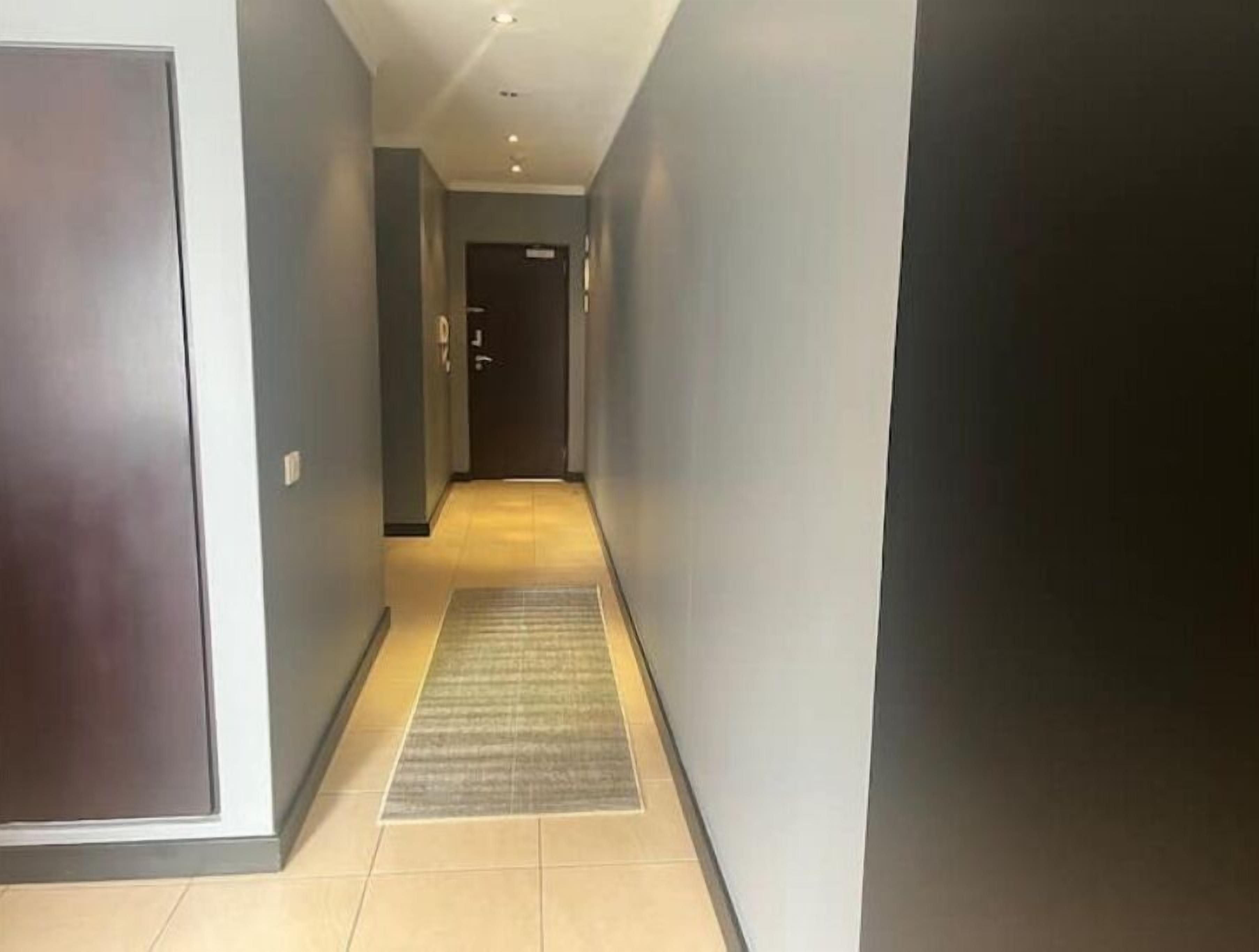 A narrow hallway with a dark door at the end, a light-colored floor, and a rug in the center.