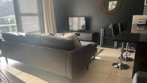 Een moderne woonkamer met een grijze bank, een televisie, een eethoek met stoelen en een groot raam.