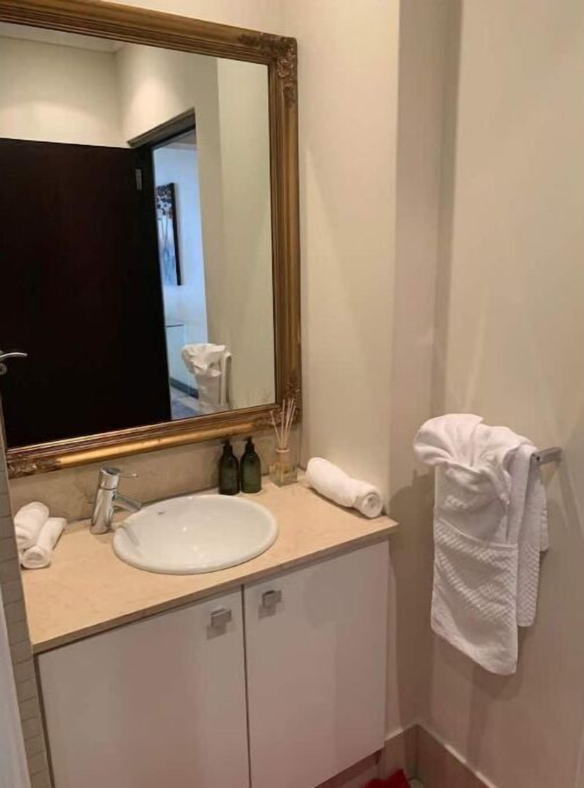 A bathroom with a white sink, a large mirror with a gold frame, a towel rack with a white towel, and a bottle of hand soap.