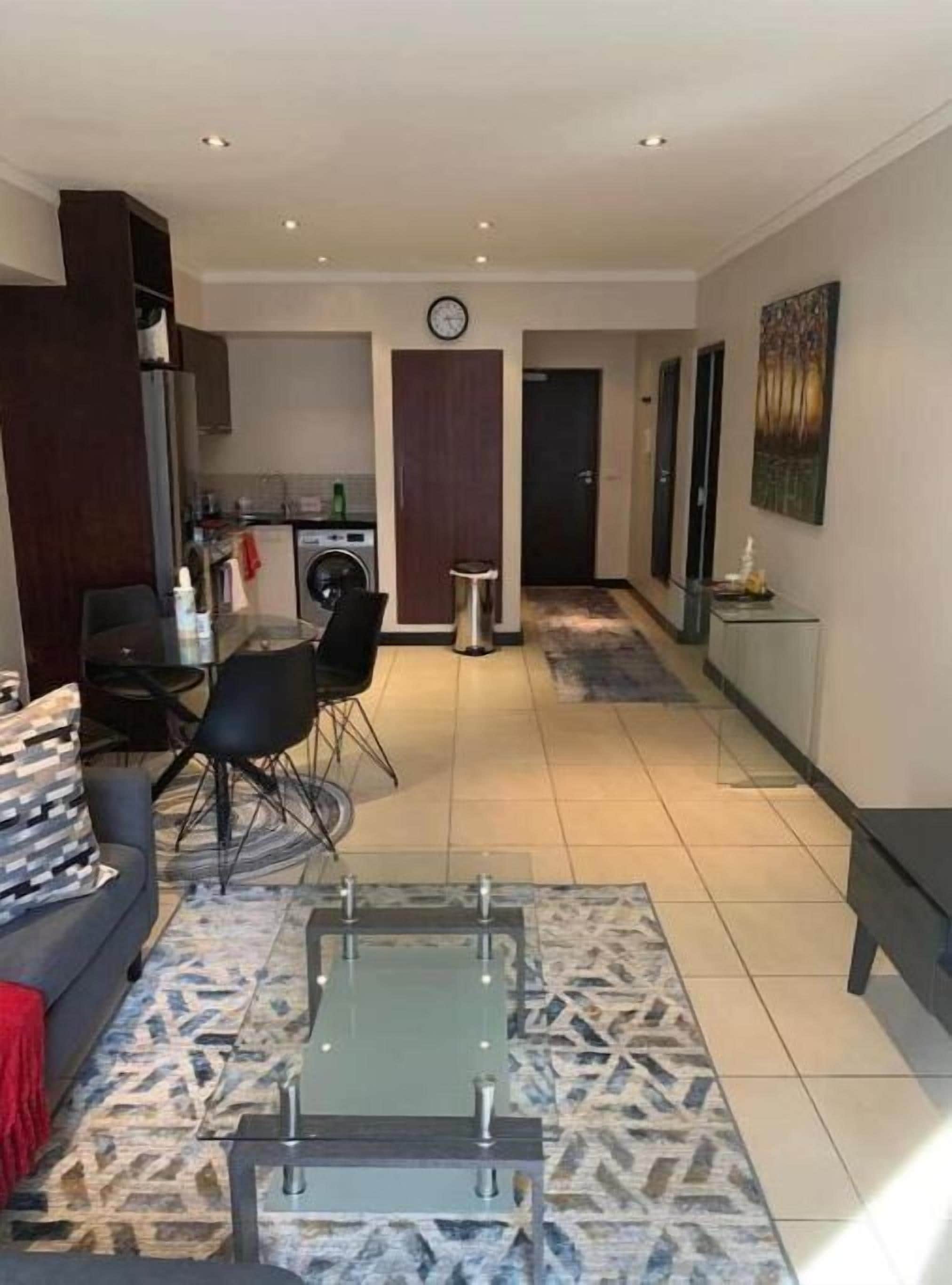 A modern living room with a sofa, coffee table, and dining area. There is a kitchen in the background with a washing machine and cabinets.