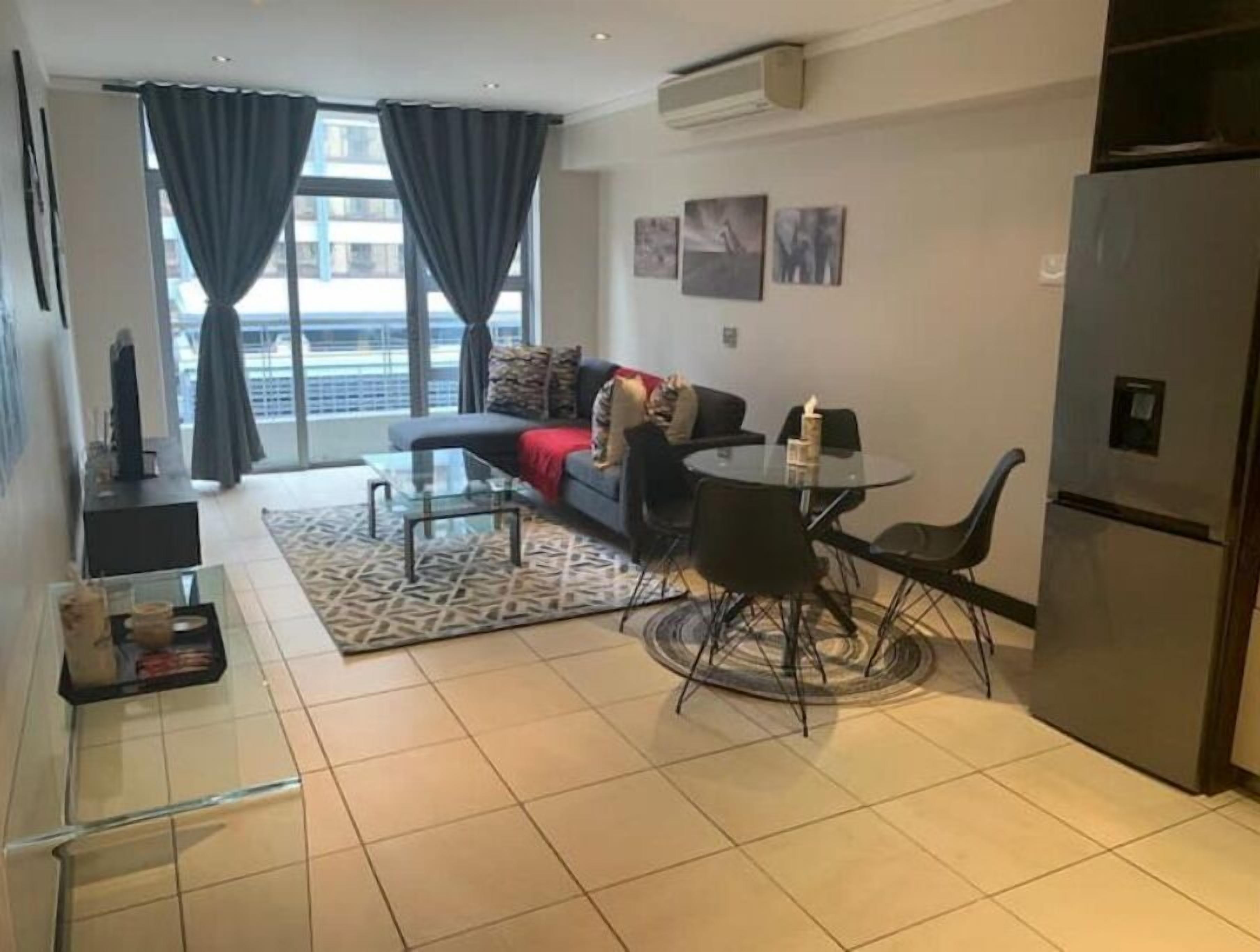 Ein modernes Wohnzimmer mit einer Couch, einem Glastisch und einem Esstisch mit Stühlen. Ein Kühlschrank, ein Fernseher und ein Fenster mit Vorhängen sind ebenfalls vorhanden.
