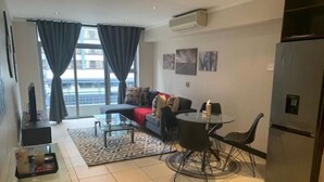 A modern living room with a sofa, a glass coffee table, and a dining table with chairs. There is a refrigerator, a TV, and a window with curtains. - Lovely Relaxing-1 Bedroon 703 Condo (Cape Town)