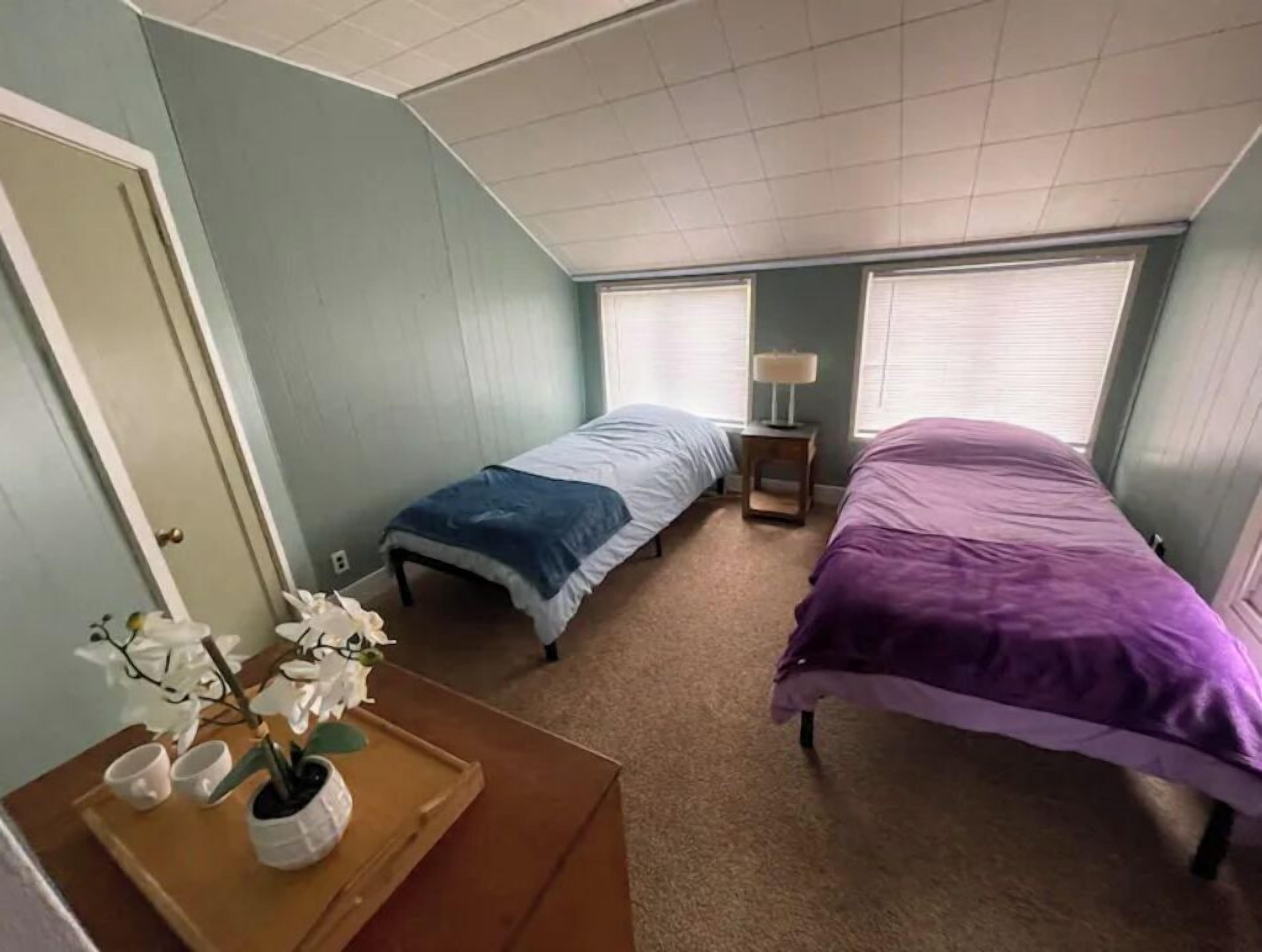 A small, single-bedroom room with two beds, a wooden dresser, a small table with a lamp, and a potted plant.