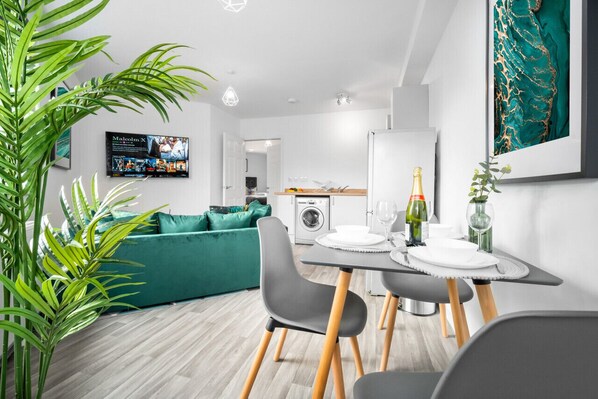 A modern living room with a green sofa, a dining table set for two, and a kitchen in the background.
