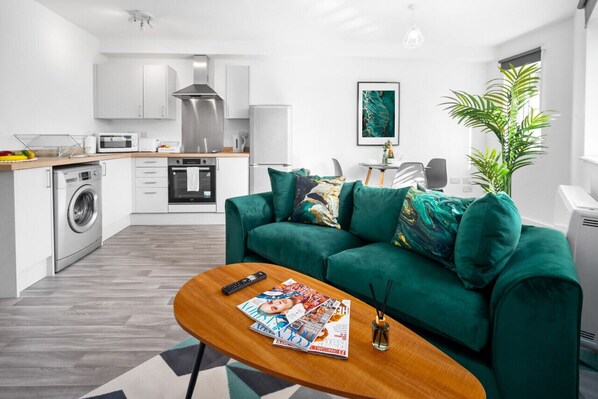 Un salon moderne avec un canapé vert, une table basse en bois sur laquelle sont posés des magazines, un lave-linge et un coin cuisine doté de meubles blancs et d’appareils électroménagers.