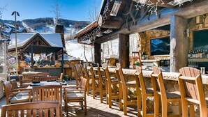 An outdoor seating area with wooden tables and chairs, a bar with bottles, and a stone wall. - Beaver Creek Luxury 2BR, 2Bath ski-in & ski-out suite inside the Ritz-Carlton (Beaver Creek)