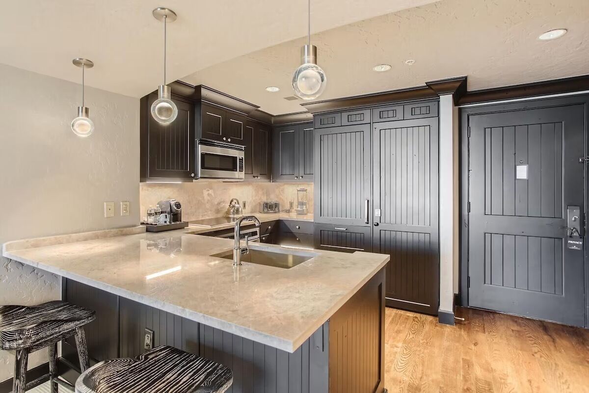 Een moderne keuken met donkere kastjes, een groot eiland met een spoelbak, roestvrijstalen apparatuur en hangende hanglampen.