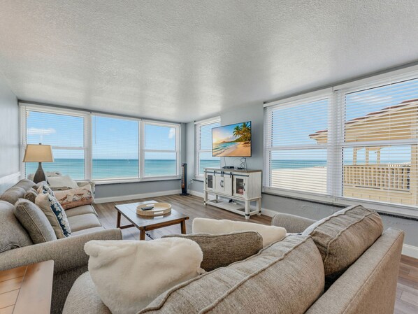 A spacious living room with a large window overlooking a beach, a flat-screen TV, and a cozy seating area.