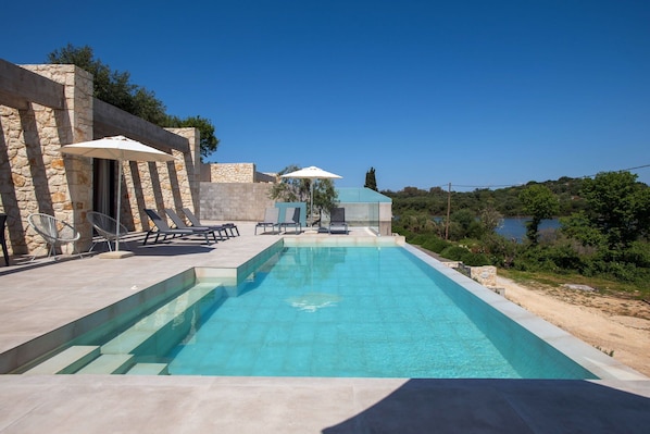 Relax by the serene pool surrounded by lush greenery and a tranquil body of water.