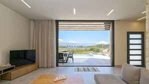 A modern living room with a TV, sofa, and coffee table, a sliding glass door to a patio with outdoor furniture, and a view of a lake and mountains.