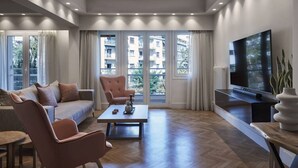 En moderne stue med sofa, stole, sofabord, fjernsyn og store vinduer med udsigt til bygninger.