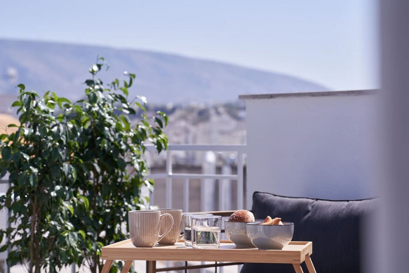 A balcony with a table set for a meal, including a bowl of bread and a glass of water, and a view of a mountain in the background.