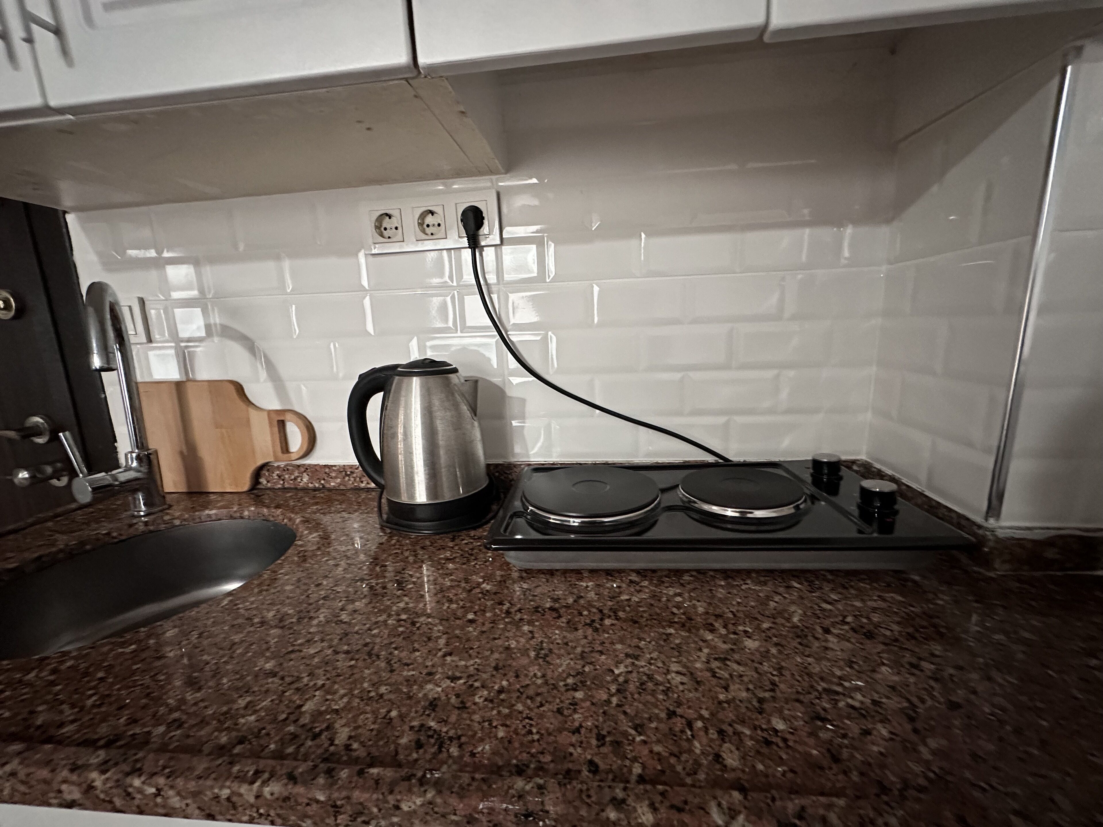 A kitchen with a granite countertop, a kettle, a stove, and a wooden cutting board.