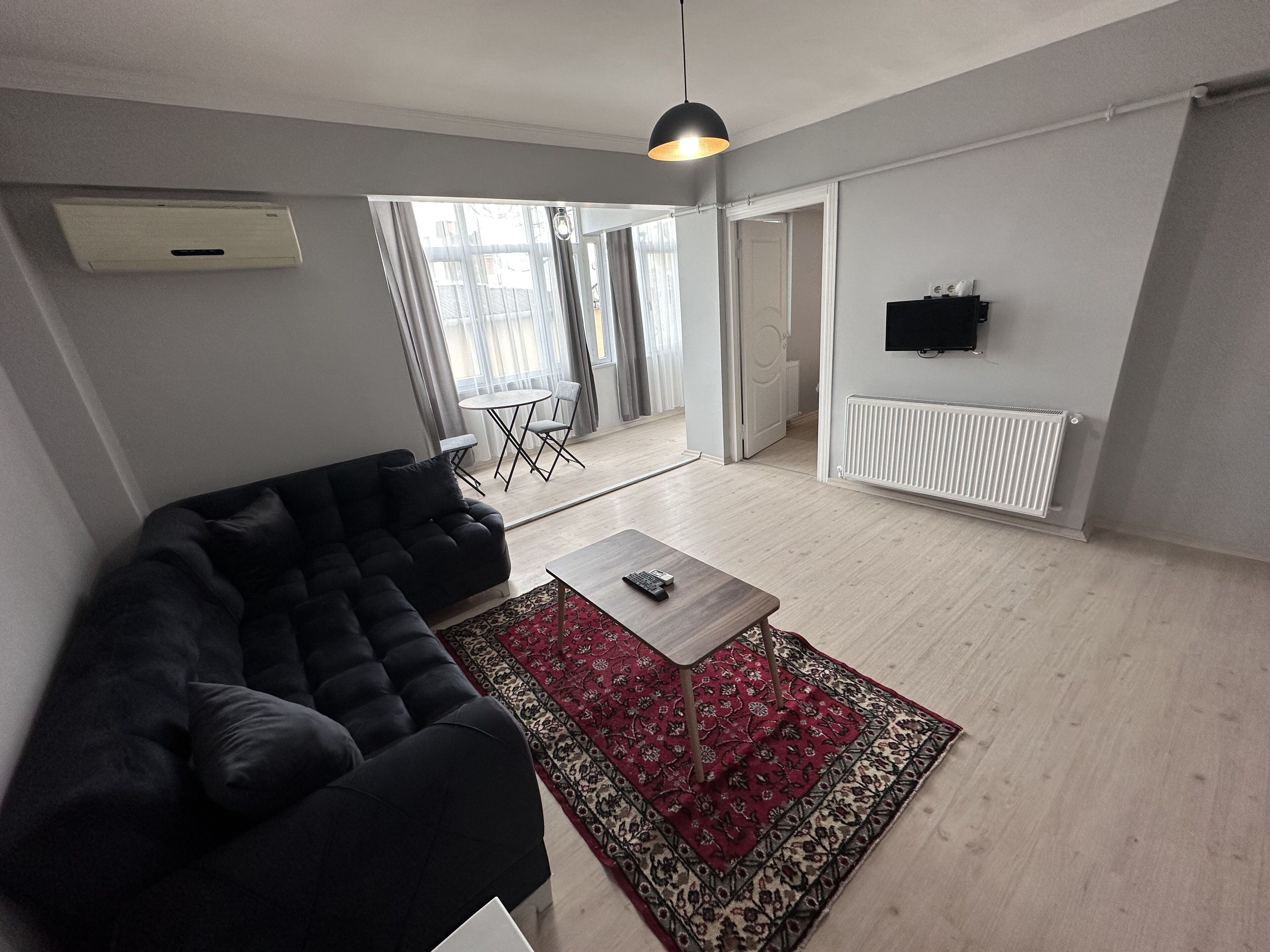 A modern living room with a black sofa, a coffee table, and a red patterned rug. There is a television mounted on the wall and a window with curtains.