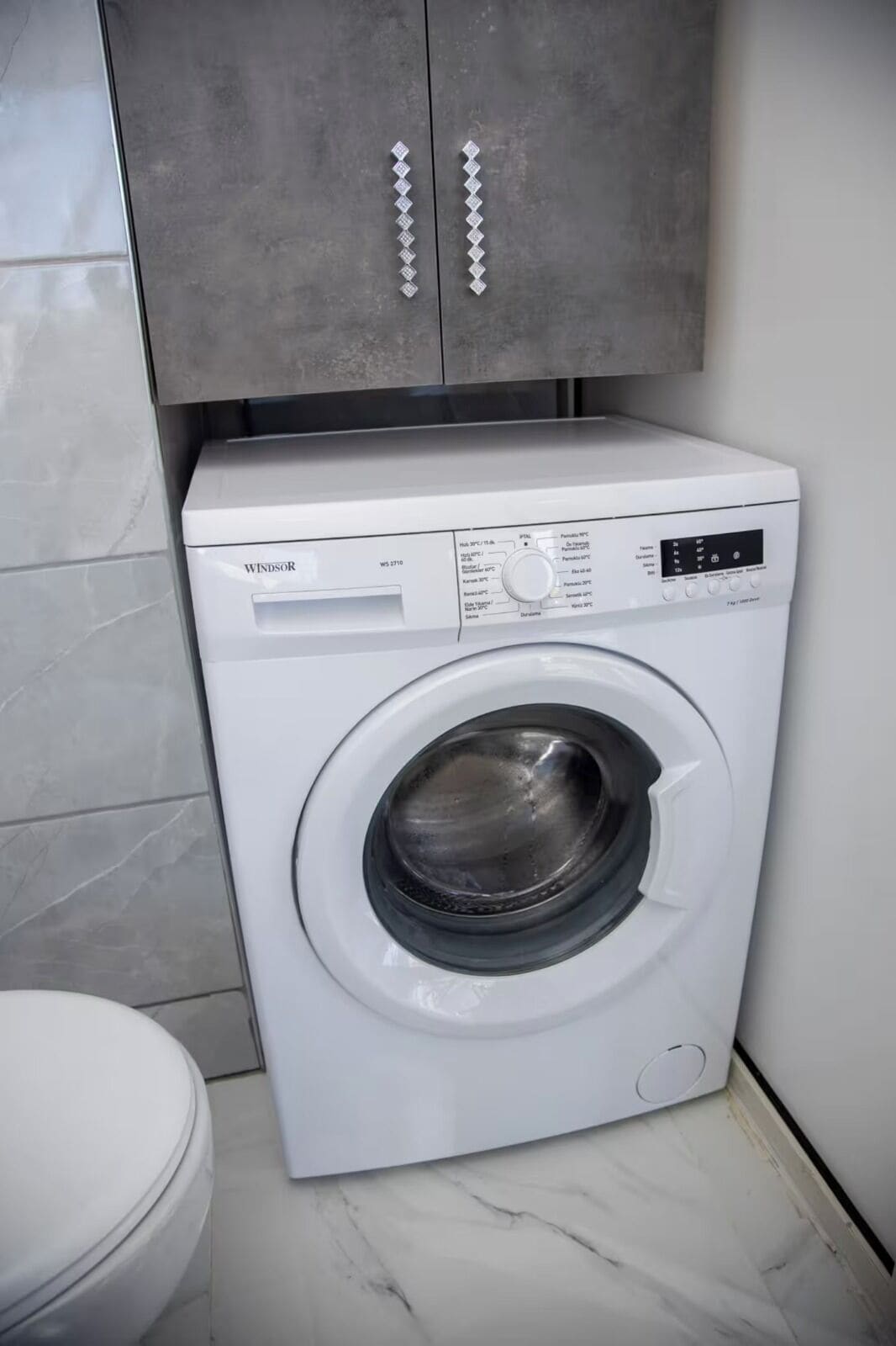 A white washing machine with a control panel, located in a bathroom with a marble floor and a grey cabinet above it.