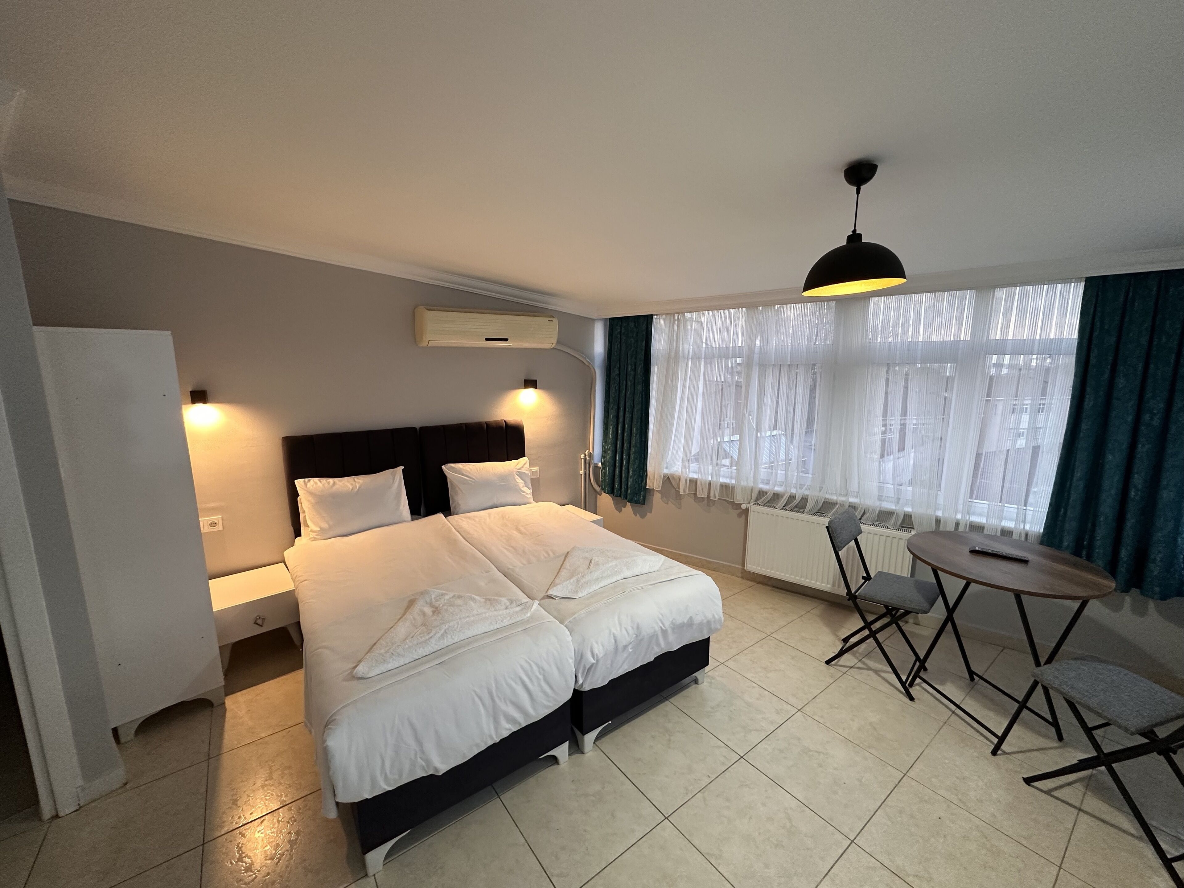 A hotel room with a large bed, bedside tables, a desk with chairs, and a window with sheer curtains.