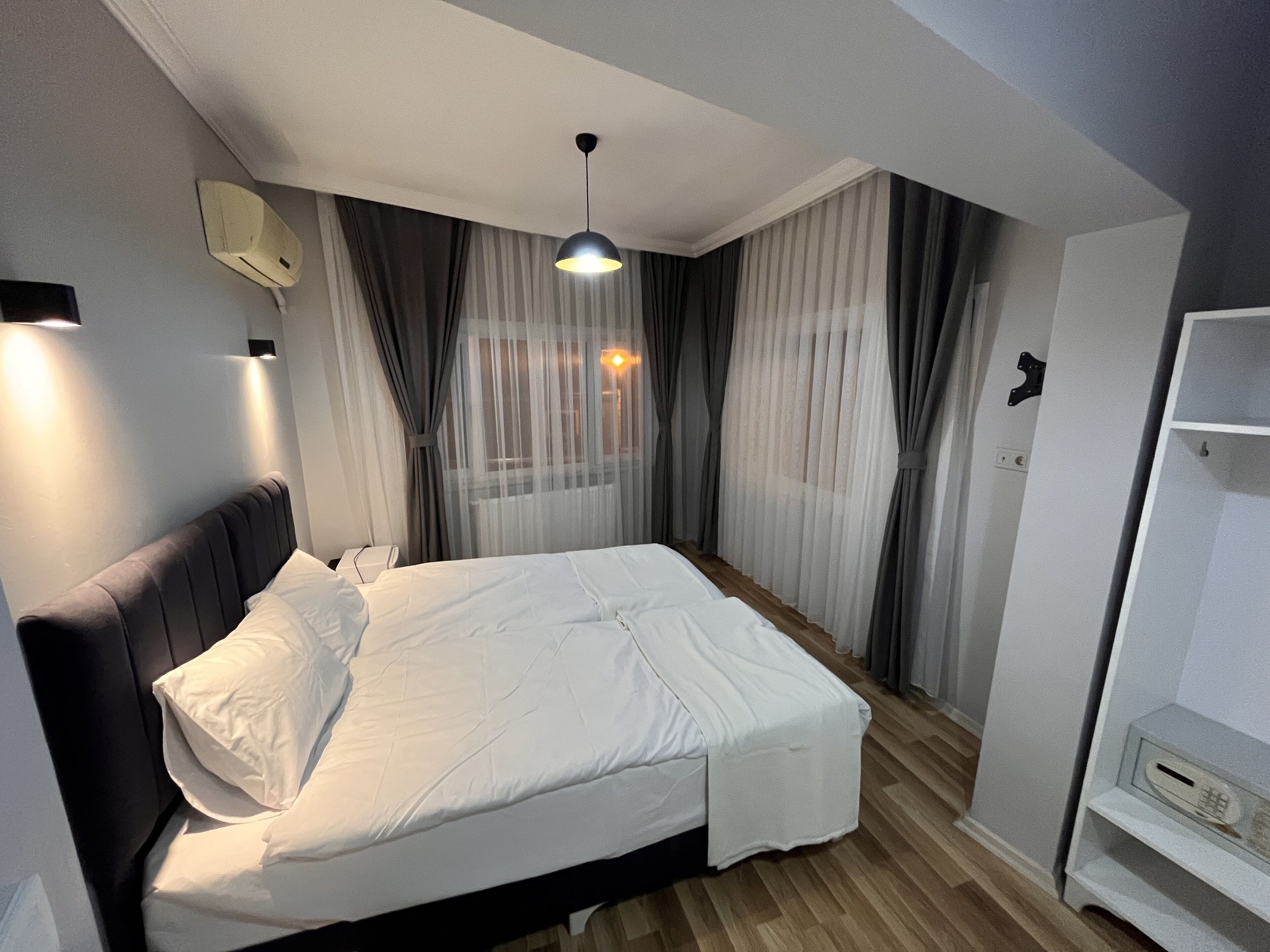 Una cama bien hecha con sábanas blancas en una habitación con suelo de madera, una luz de techo y cortinas traslúcidas.