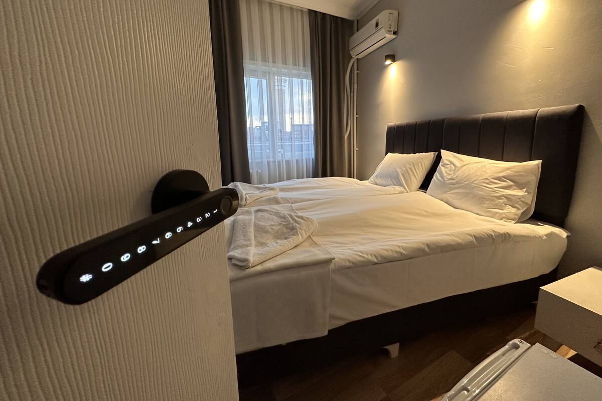 A hotel room with a bed, a bedside table, an air conditioning unit, and a window with curtains.