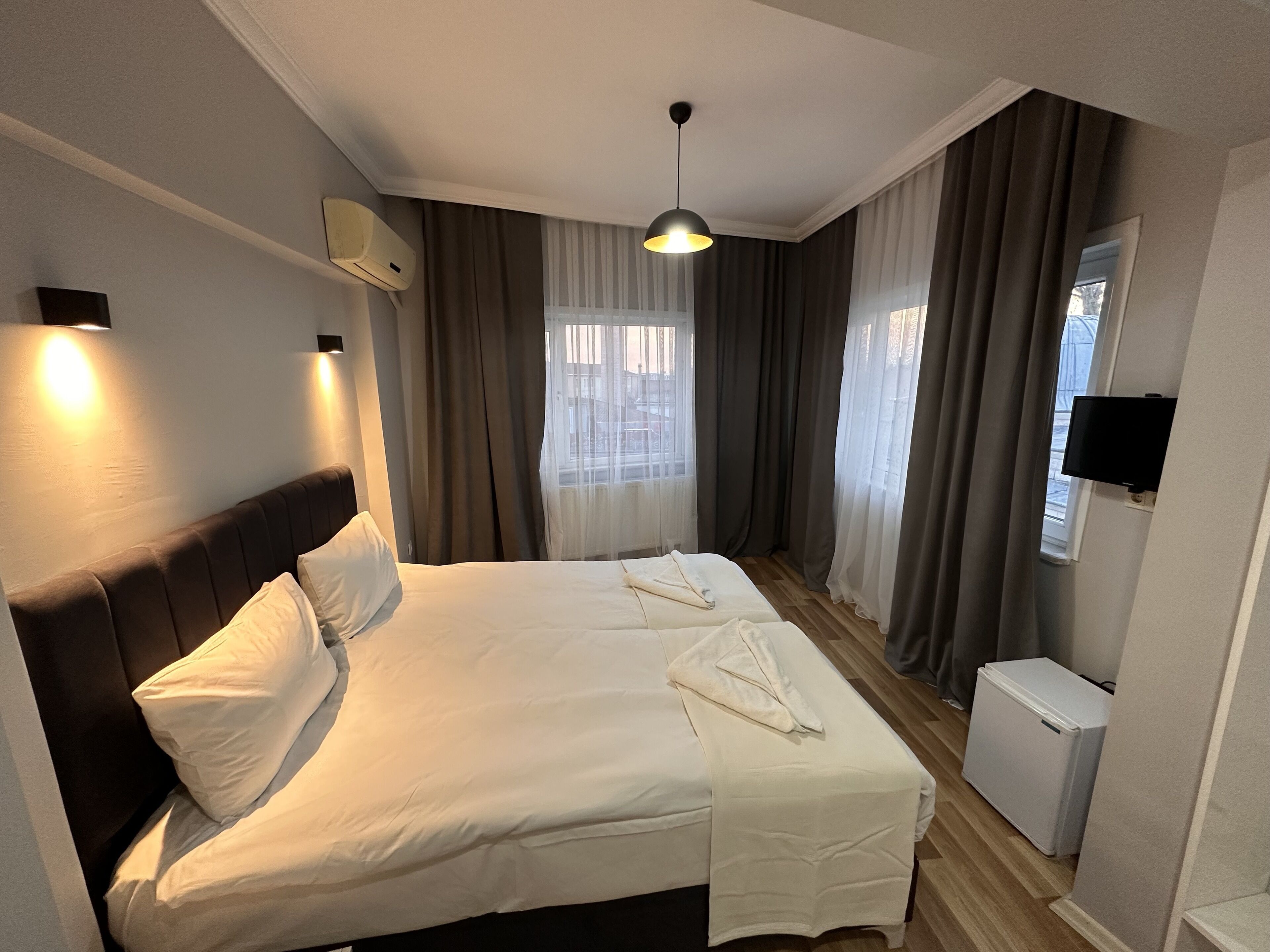 A hotel room with a large bed, a flat-screen TV, an air conditioning unit, and a window with curtains.