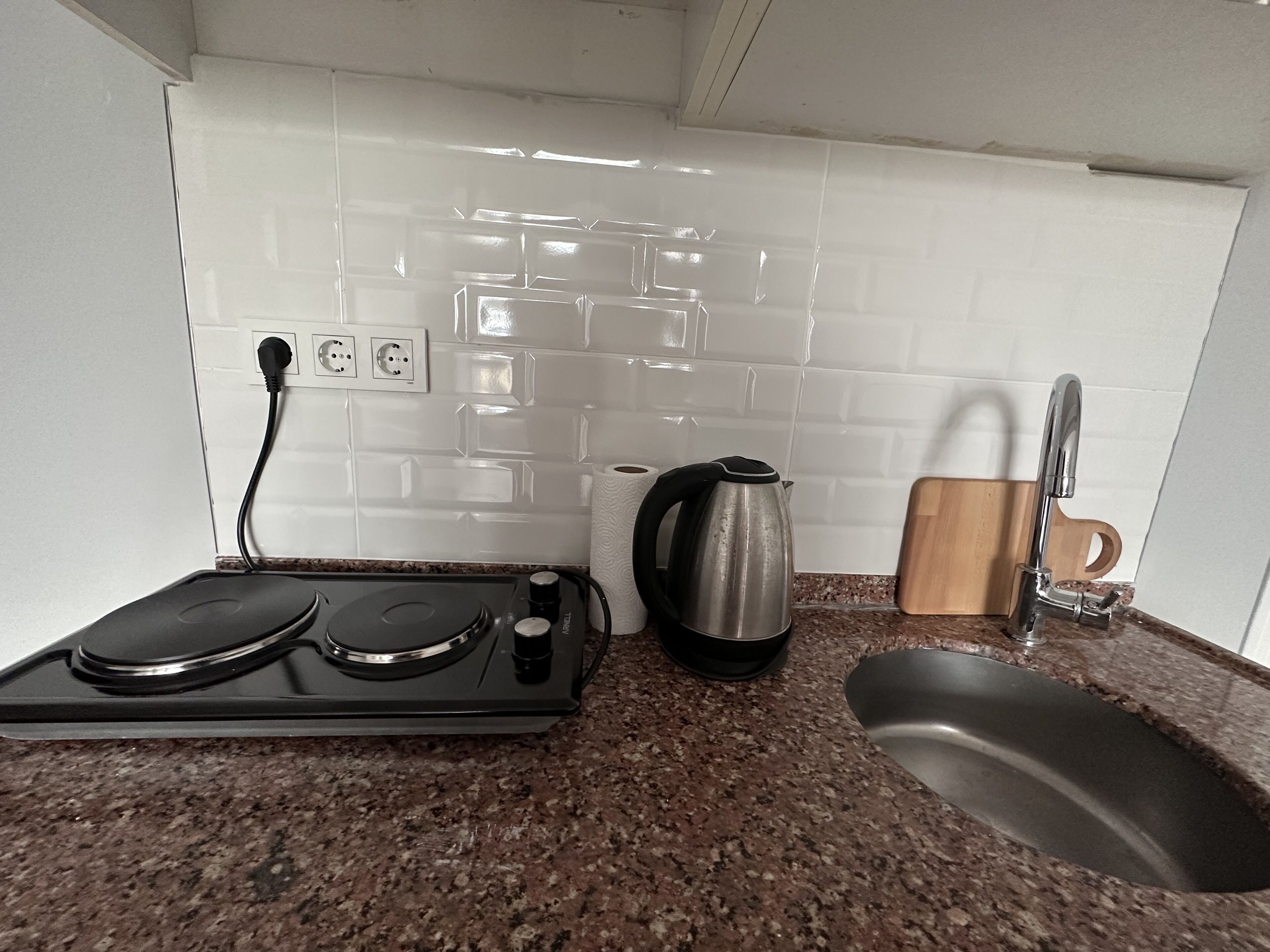 A kitchen with a gas stove, a kettle, a sink, and a wooden cutting board.