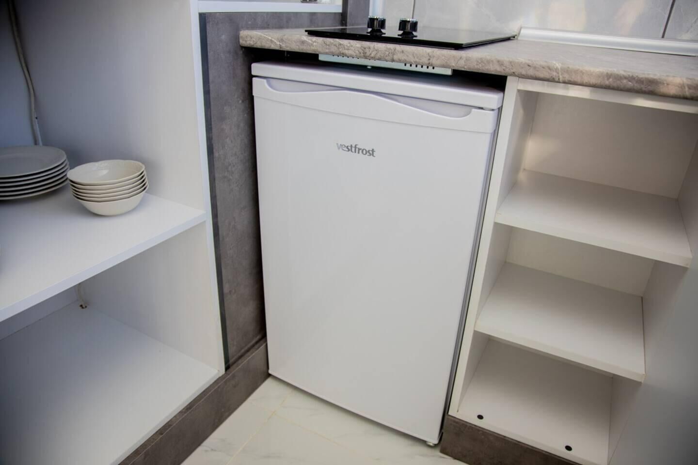 A compact kitchen with a white dishwasher, a countertop with a stove, and a stack of plates on a shelf.