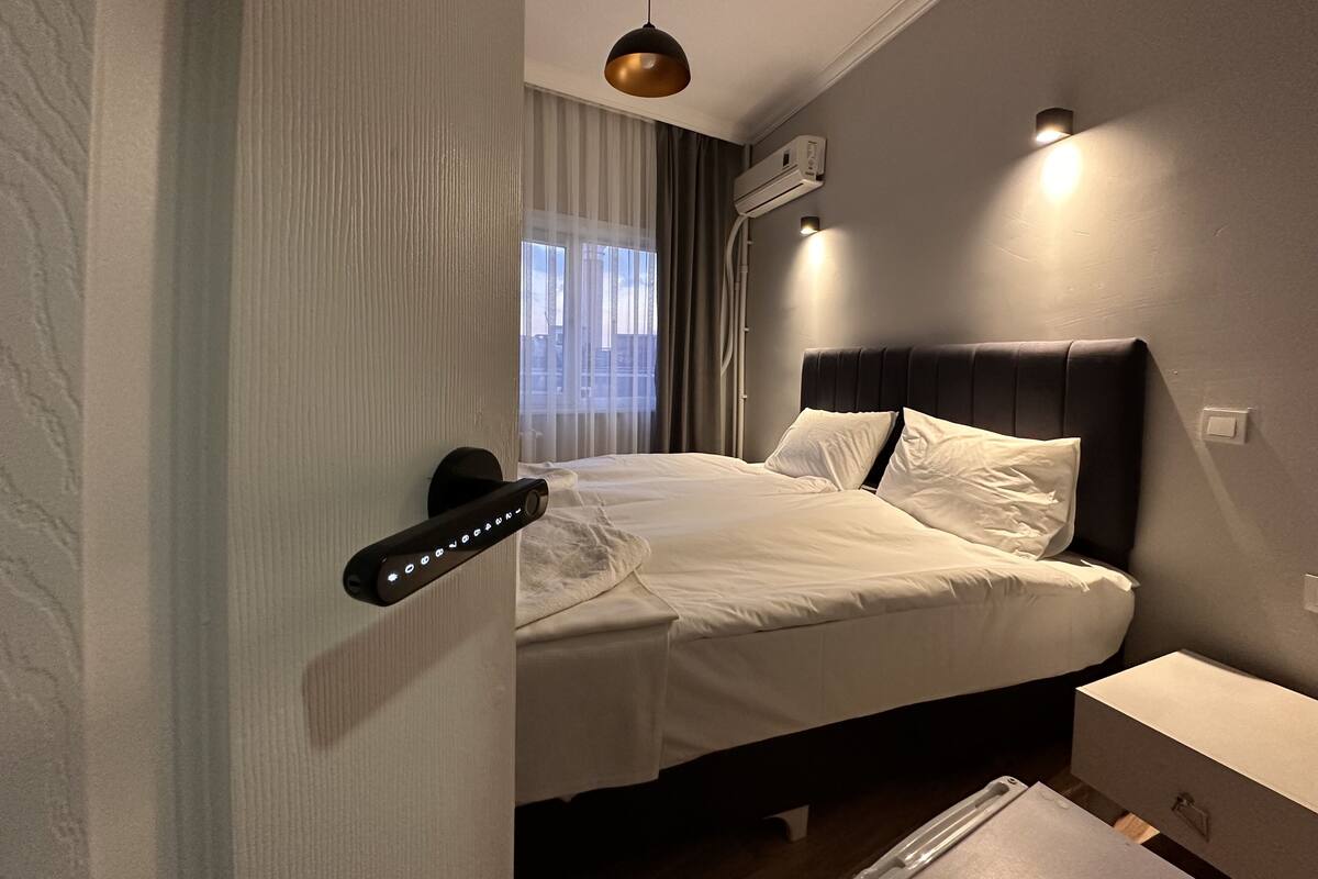 A modern hotel room with a bed, bedside table, and a control panel on the wall.