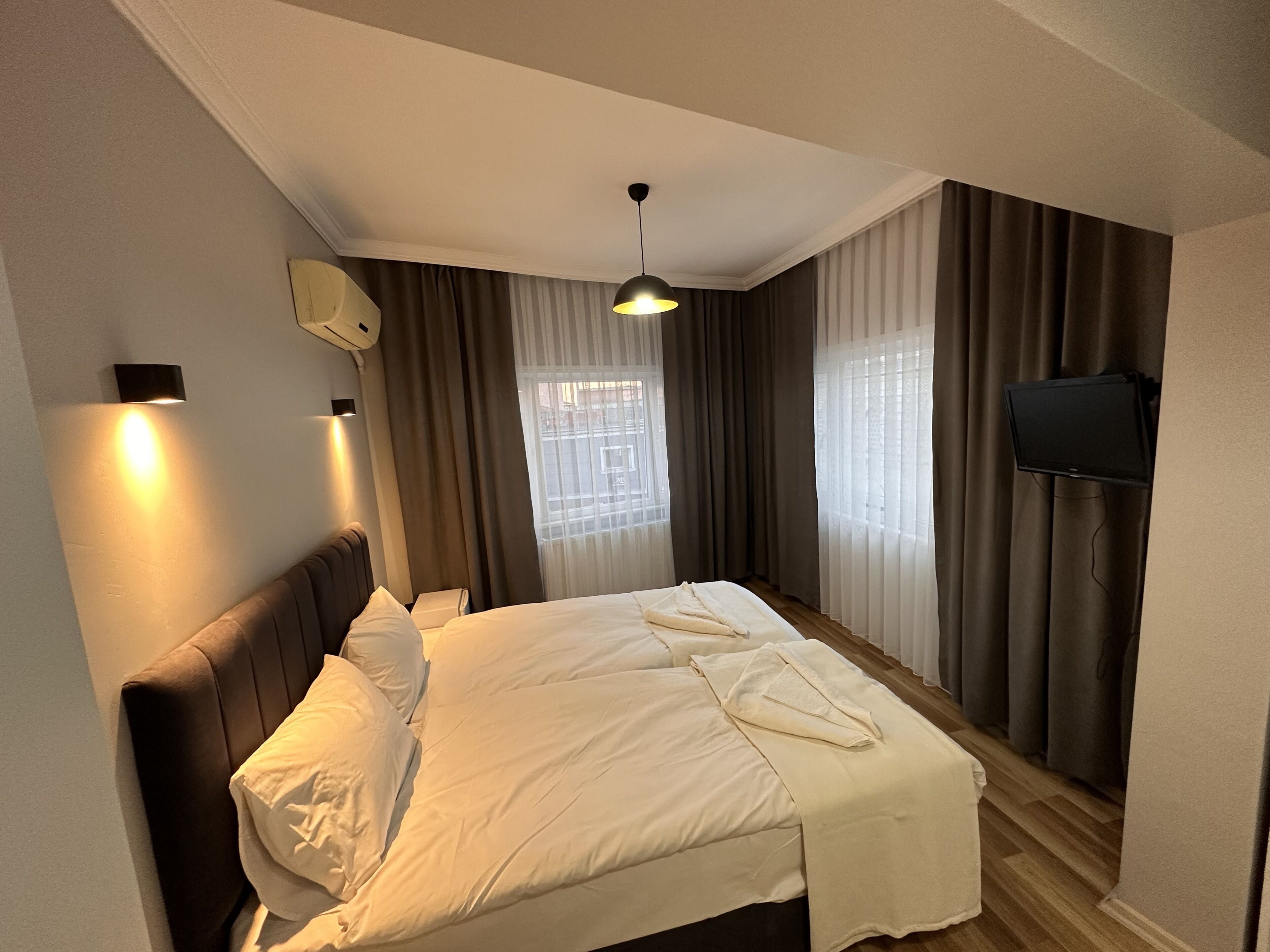 A hotel room with a bed, curtains, a mounted TV, and a ceiling light.