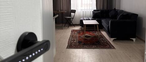 A modern living room with a black sofa, a small coffee table, and a red rug. There is a dining area with a table and chairs, and a large window with curtains.