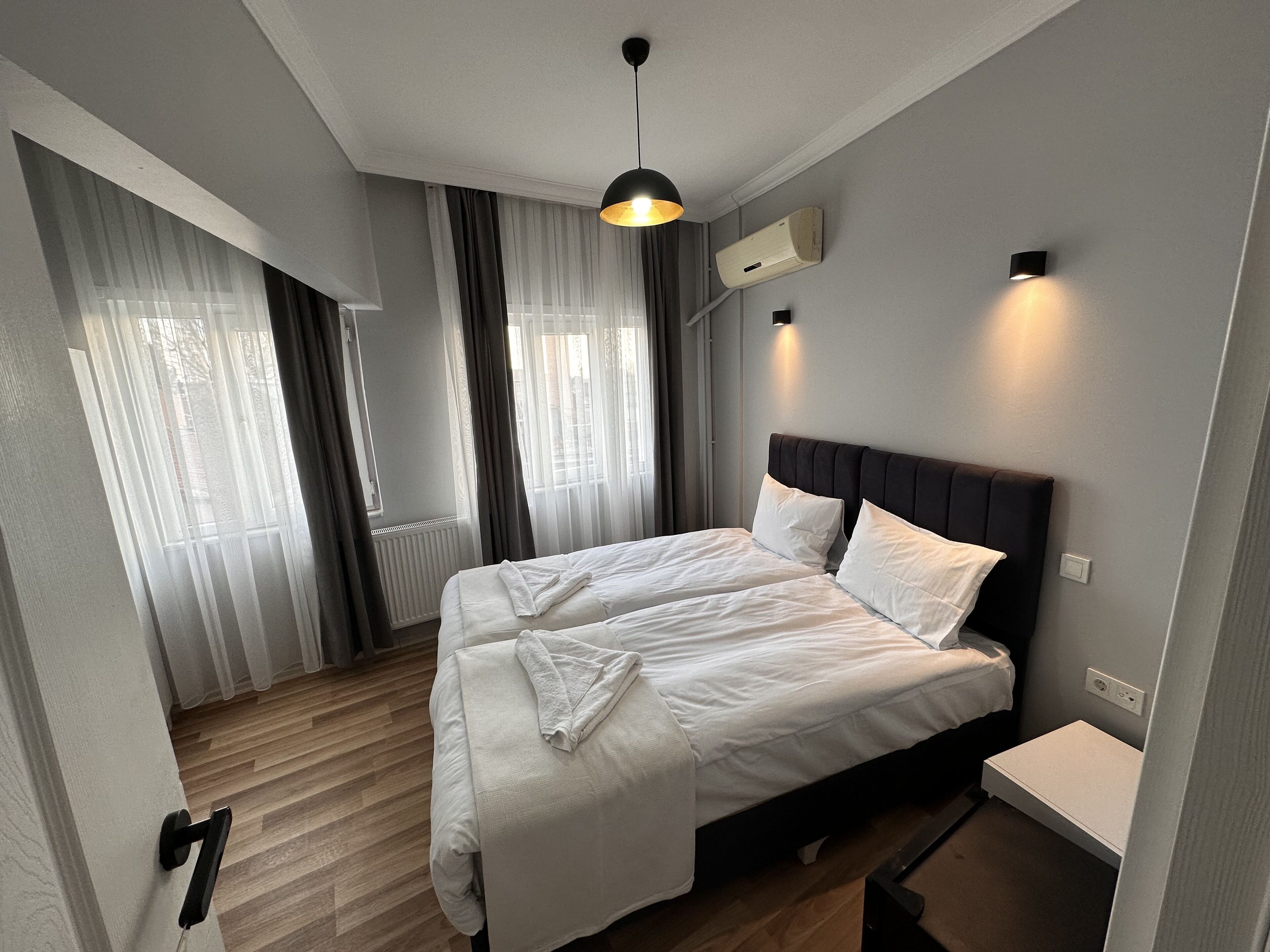 Ein modernes Schlafzimmer mit einem großen Bett, einem Nachttisch, einer Pendelleuchte und einem Fenster mit Vorhängen.