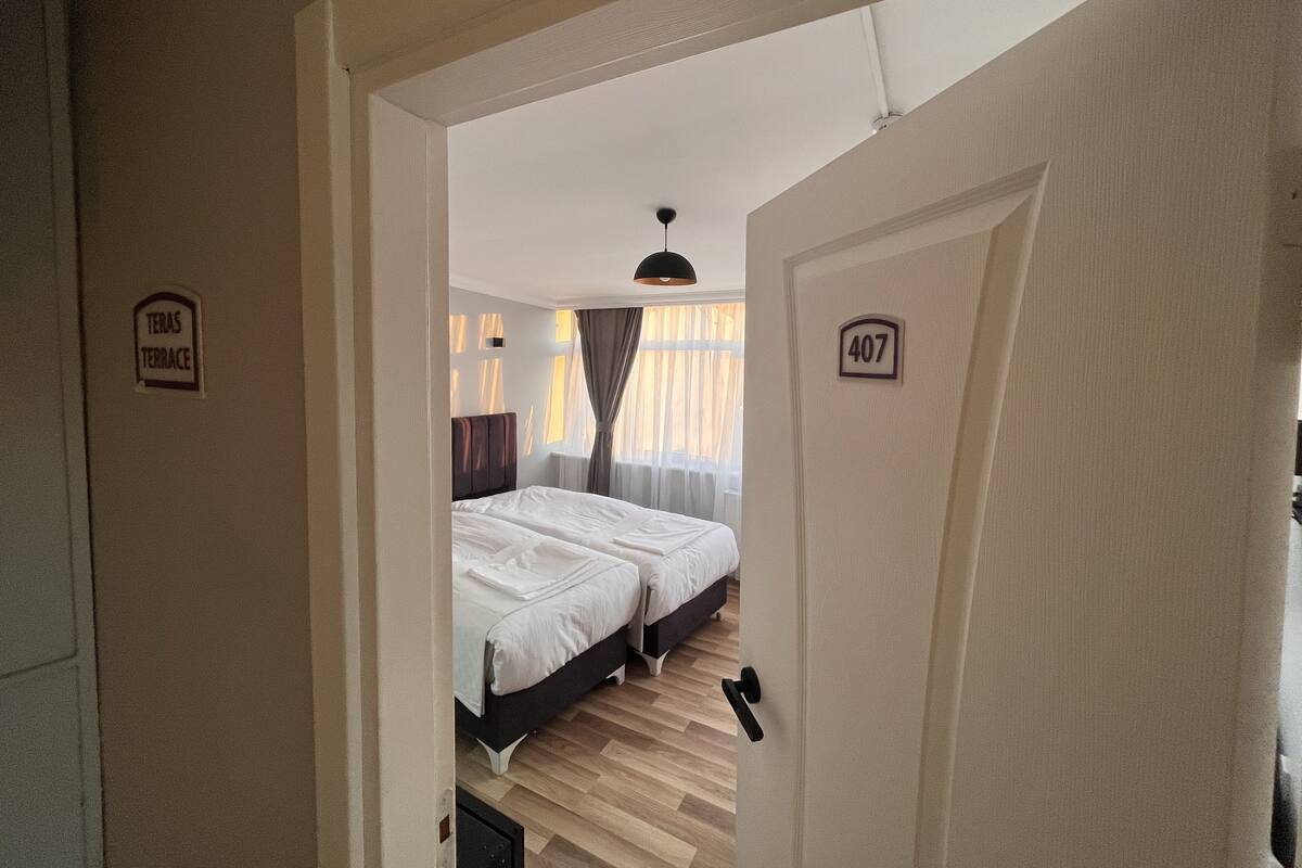 A hotel room with two beds, a wooden floor, a window with curtains, and a door with a number sign.