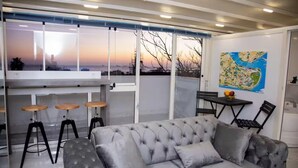 A modern living room with a grey tufted sofa, a bar area with stools, a dining table with chairs, and a large window with a view of the sunset. - Asparagus Hotel Suits Old City (Istanbul)